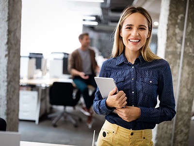 A young female wearing yellow pants and a dark blue blouse smiles while holding a tablet after seeing her BlueCross BlueShield dentist in McKinney