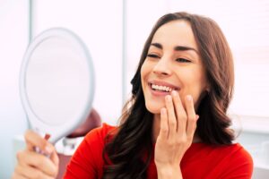 Woman with brown hair in red shirt smiling at her reflection and touching her teeth